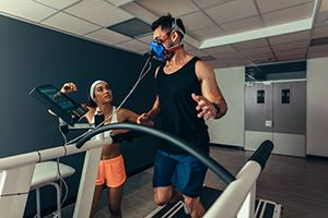 Man running on treadmill with fitness mask on while woman is looking on watching screen with results on it.